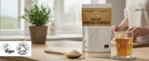 Organic Haritaki Powder packaging with a glass of drink and plant in background.